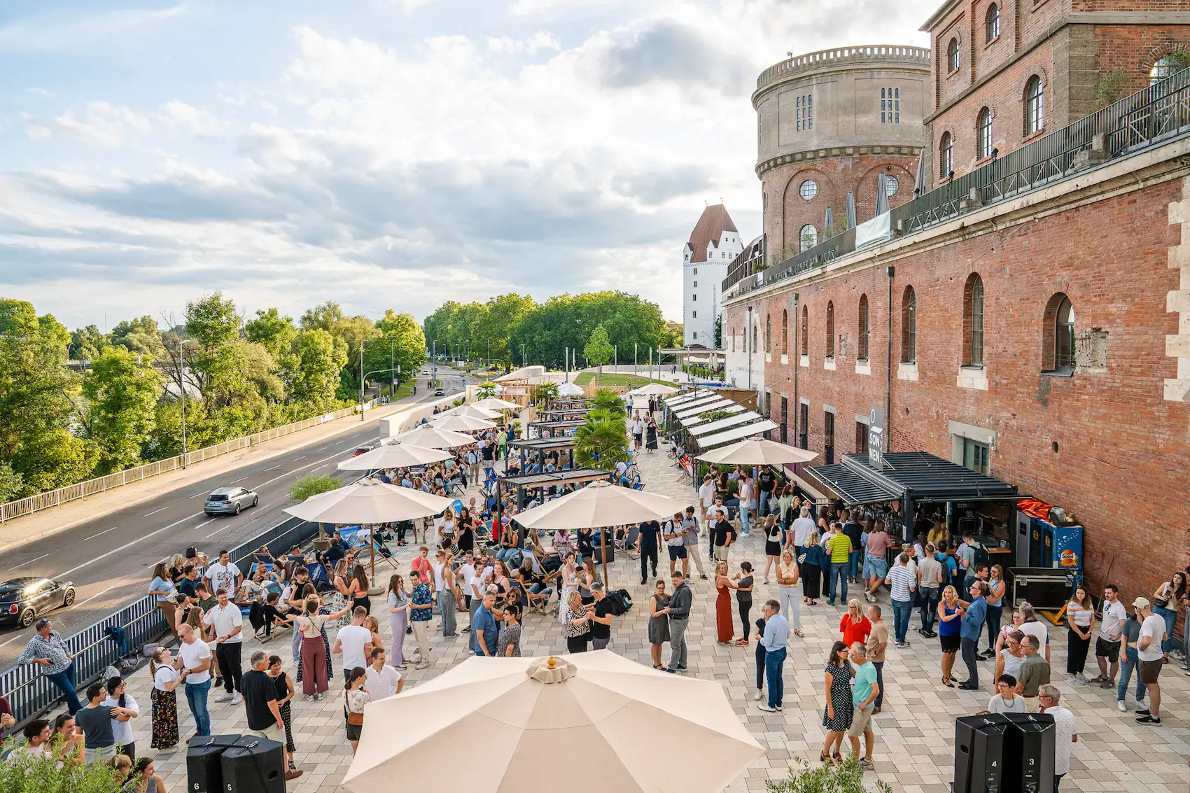 Menschen versammeln sich unter Sonnenschirmen vor einem roten Backsteingebäude an einem sonnigen Tag neben einer Straße mit grünem Baumbewuchs.
