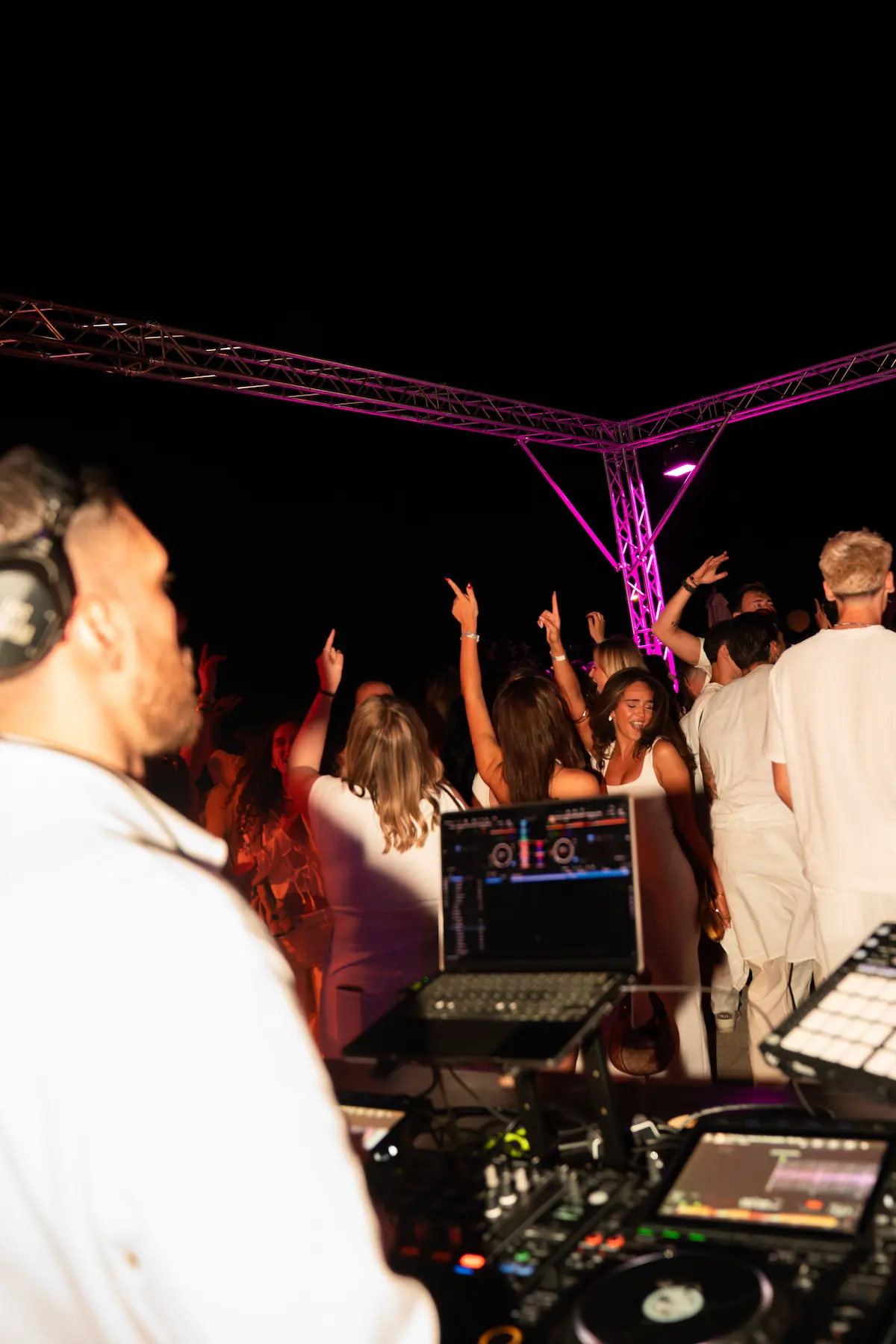 DJ mit Kopfhörern legt Musik auf einer Party mit tanzender Menge unter rosa Beleuchtung auf.
