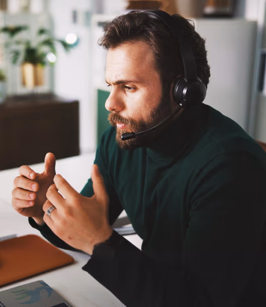 Man with beard wearing headset gesturing with hands while working at desk indoors.