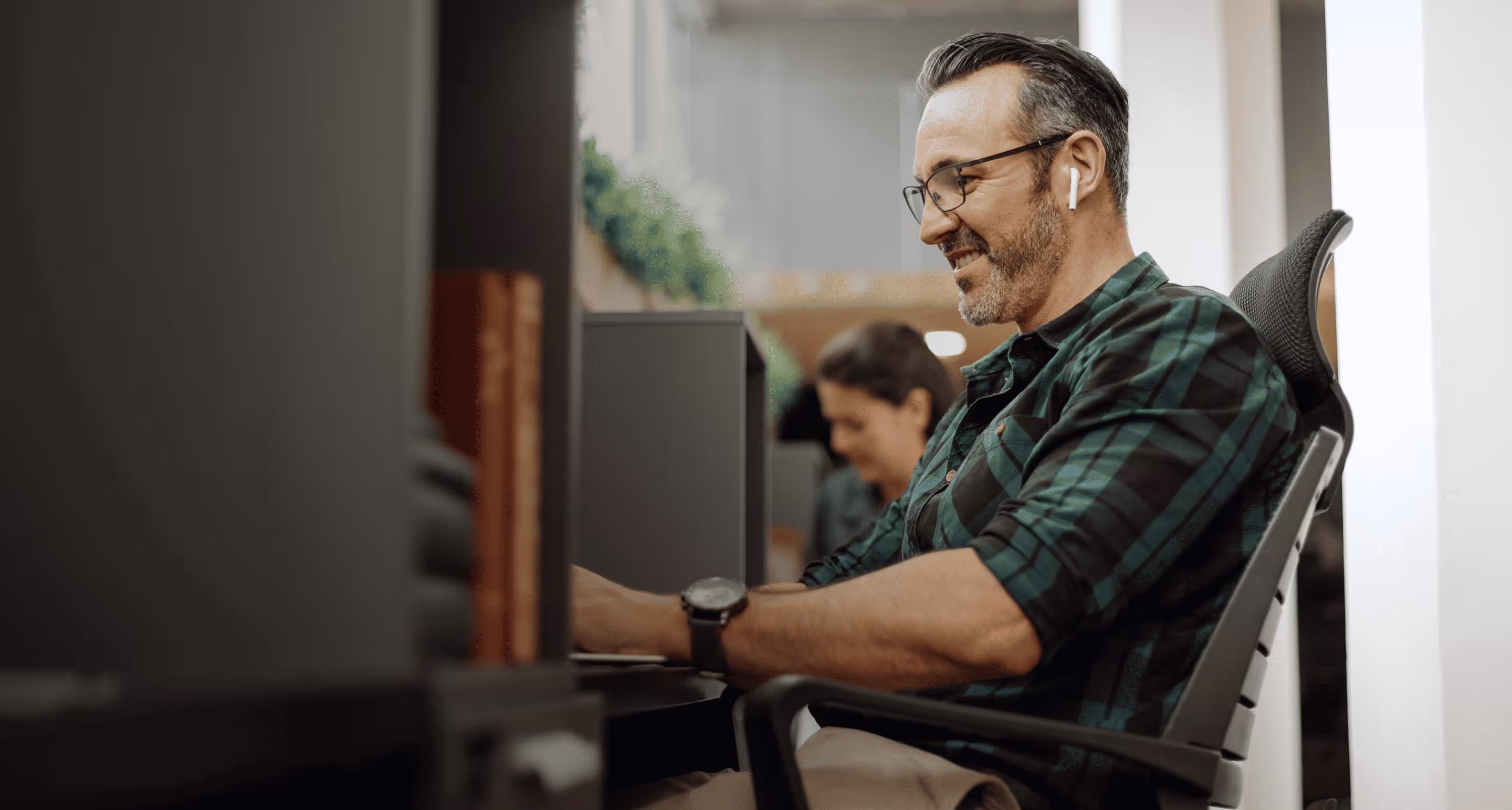 Man smiling and working on a laptop at a desk wearing a green plaid shirt and wireless earbuds.