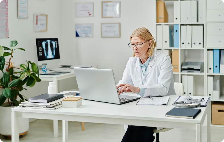 Female doctor wearing glasses working on a laptop at a desk in a modern medical office.
