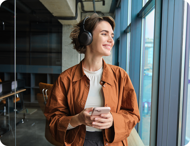 Smiling woman wearing headphones, holding a smartphone, looking out a large office window.