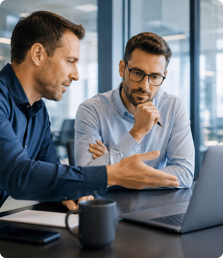 Two men in an office discussing work while looking at a laptop screen on a desk.