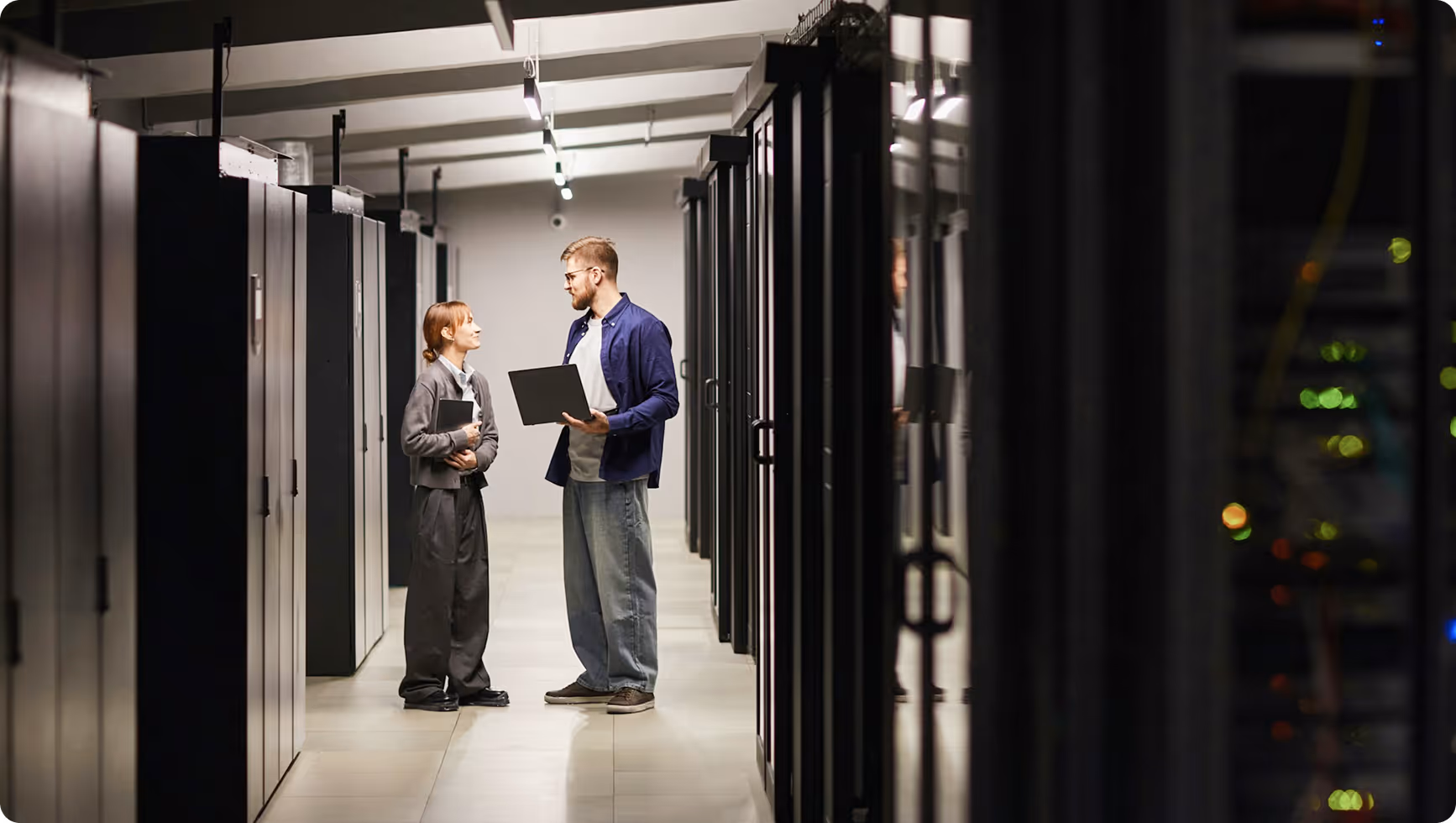 Two IT professionals standing and discussing with laptops in a server room aisle.