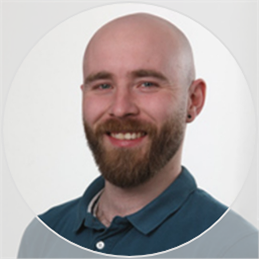 Smiling bald man with a beard wearing a dark blue collared shirt against a light background.
