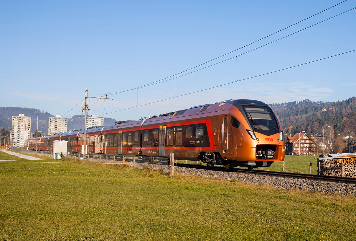 Modern orange SOB train traveling on tracks beside green field with houses and hills in background.