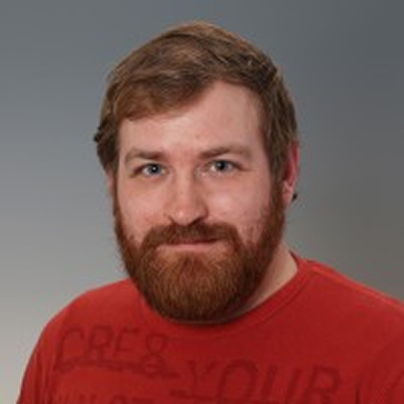 Portrait of a smiling man with red hair and beard wearing a red shirt against a gray background.
