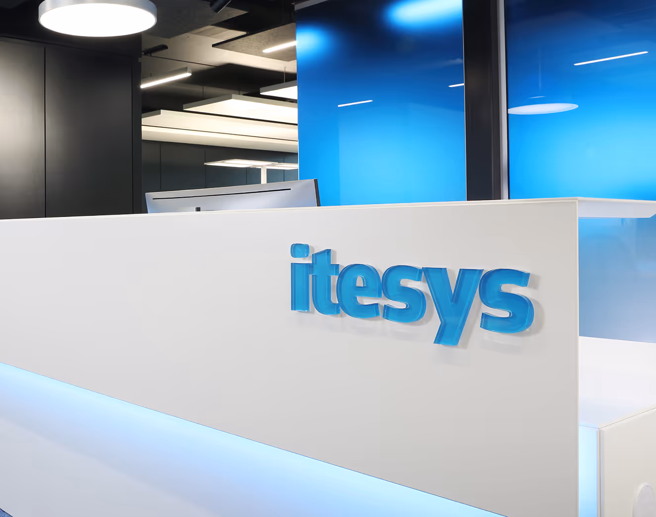 Modern office reception desk with a blue 'itesys' logo and blue-lit panel below.