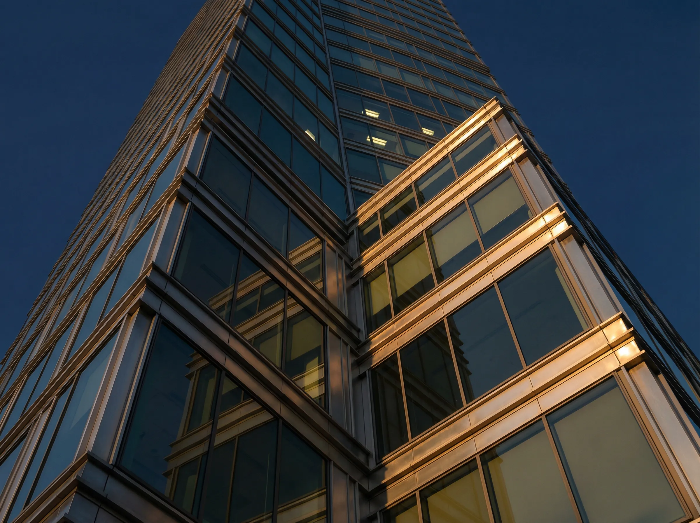 Upward view of a modern glass skyscraper corner reflecting another building during sunset.