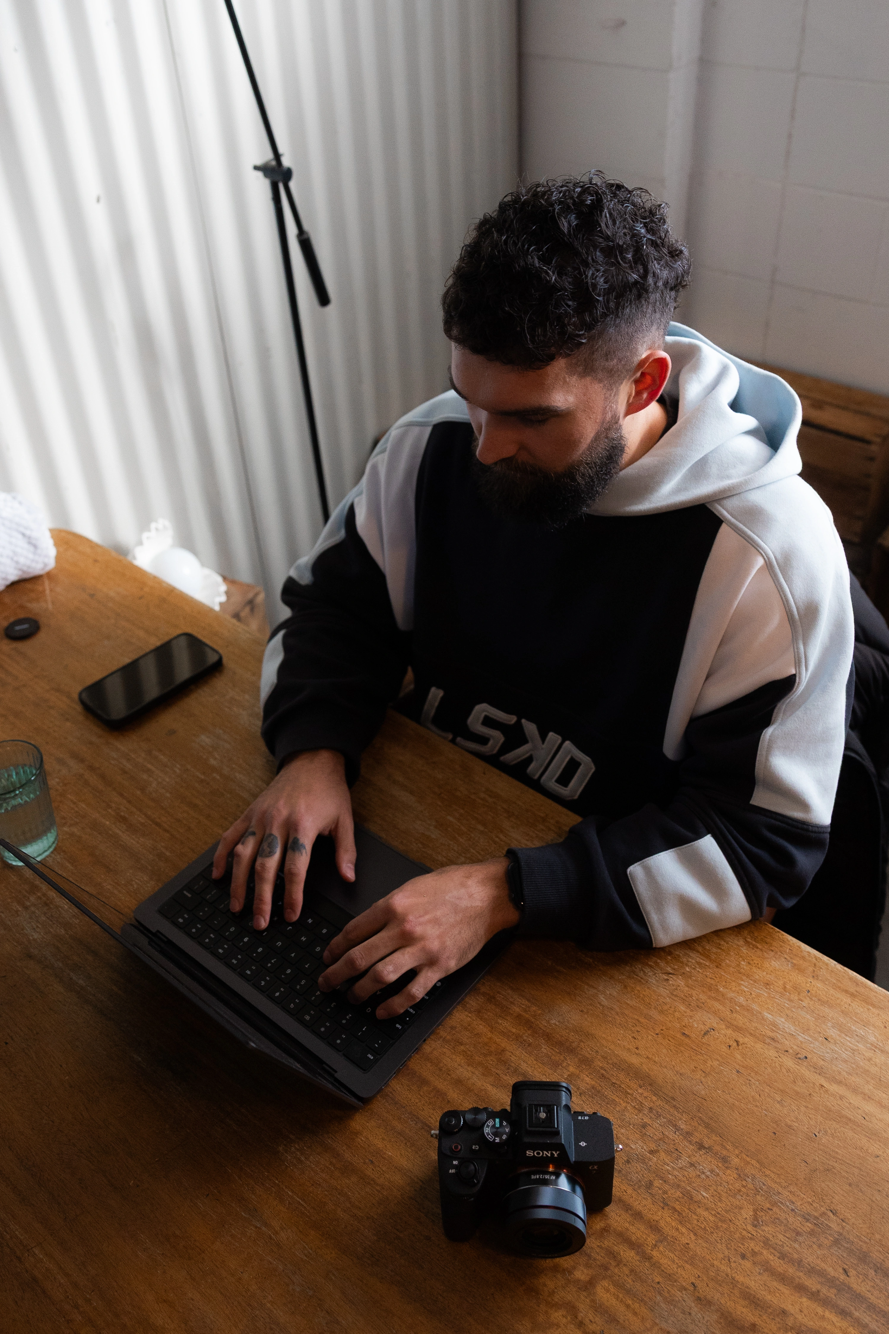 Man with curly hair and beard typing on a laptop at a wooden table with a Sony camera and smartphone nearby.
