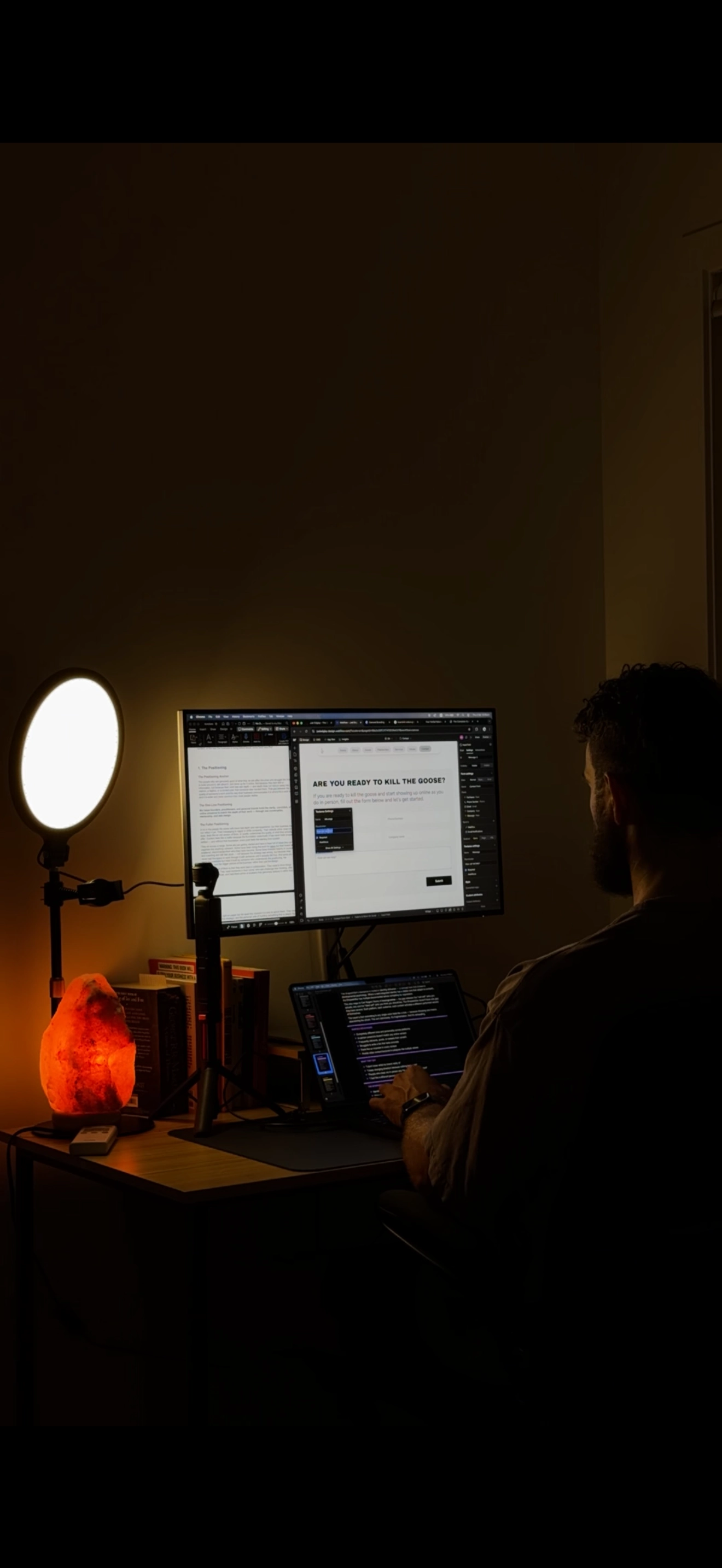 Man working at a desk in a dimly lit room with a monitor, laptop, ring light, salt lamp, and books.