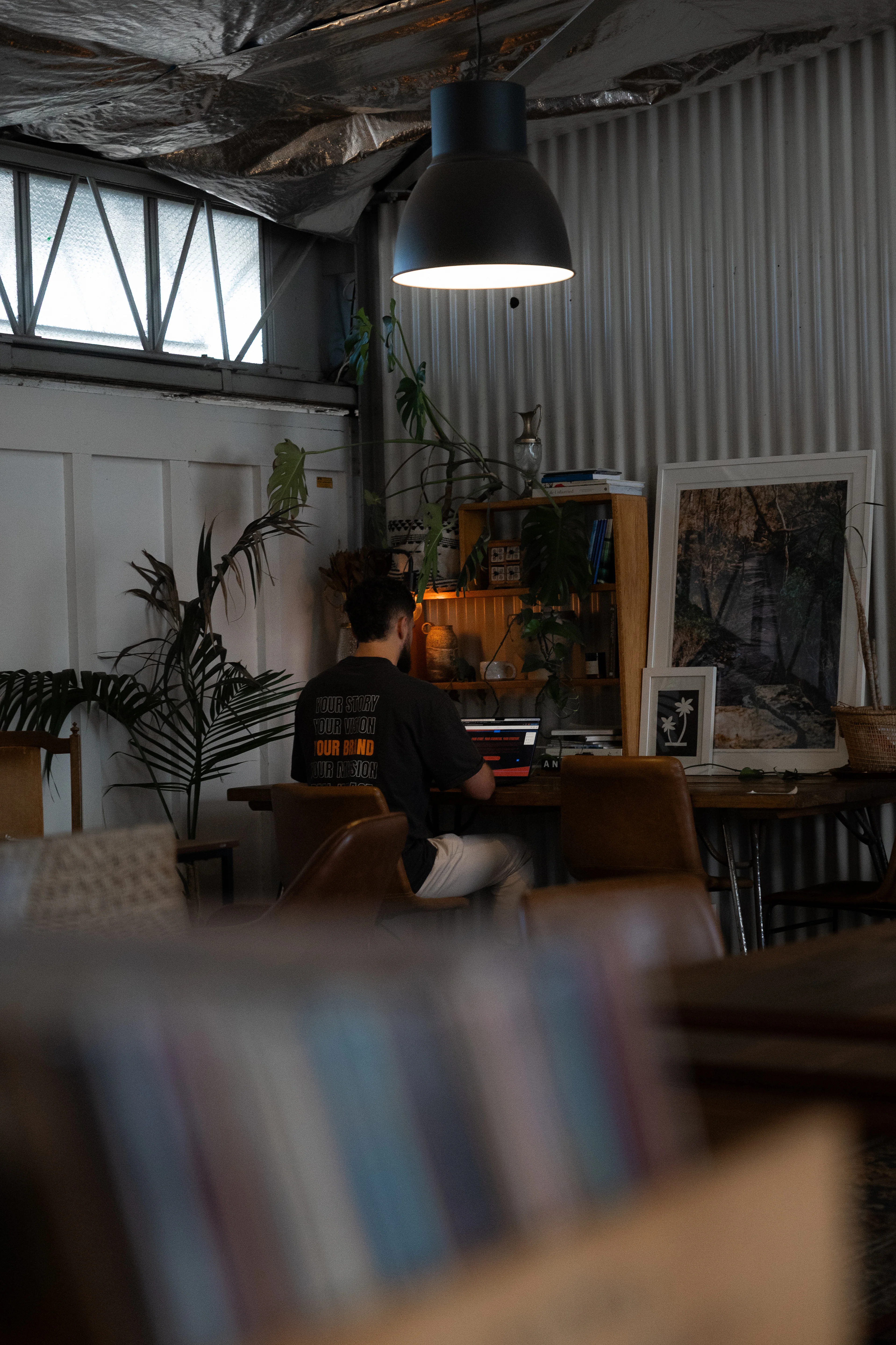 Person sitting at a desk working on a laptop under a hanging lamp in a cozy, plant-filled room with framed artwork on the wall.