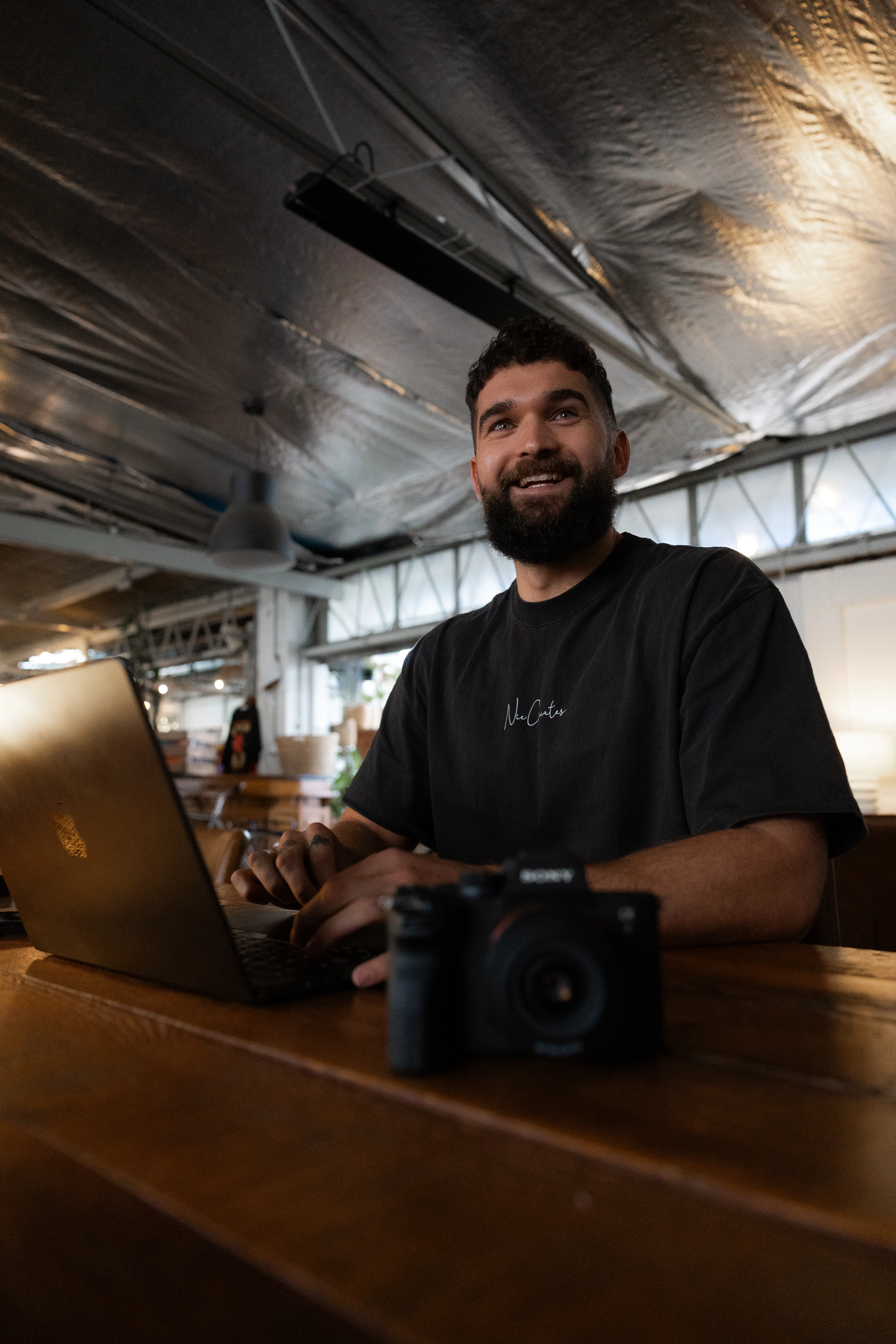 Bearded man smiling and typing on a laptop at a wooden table with a camera in front in an industrial-style room.