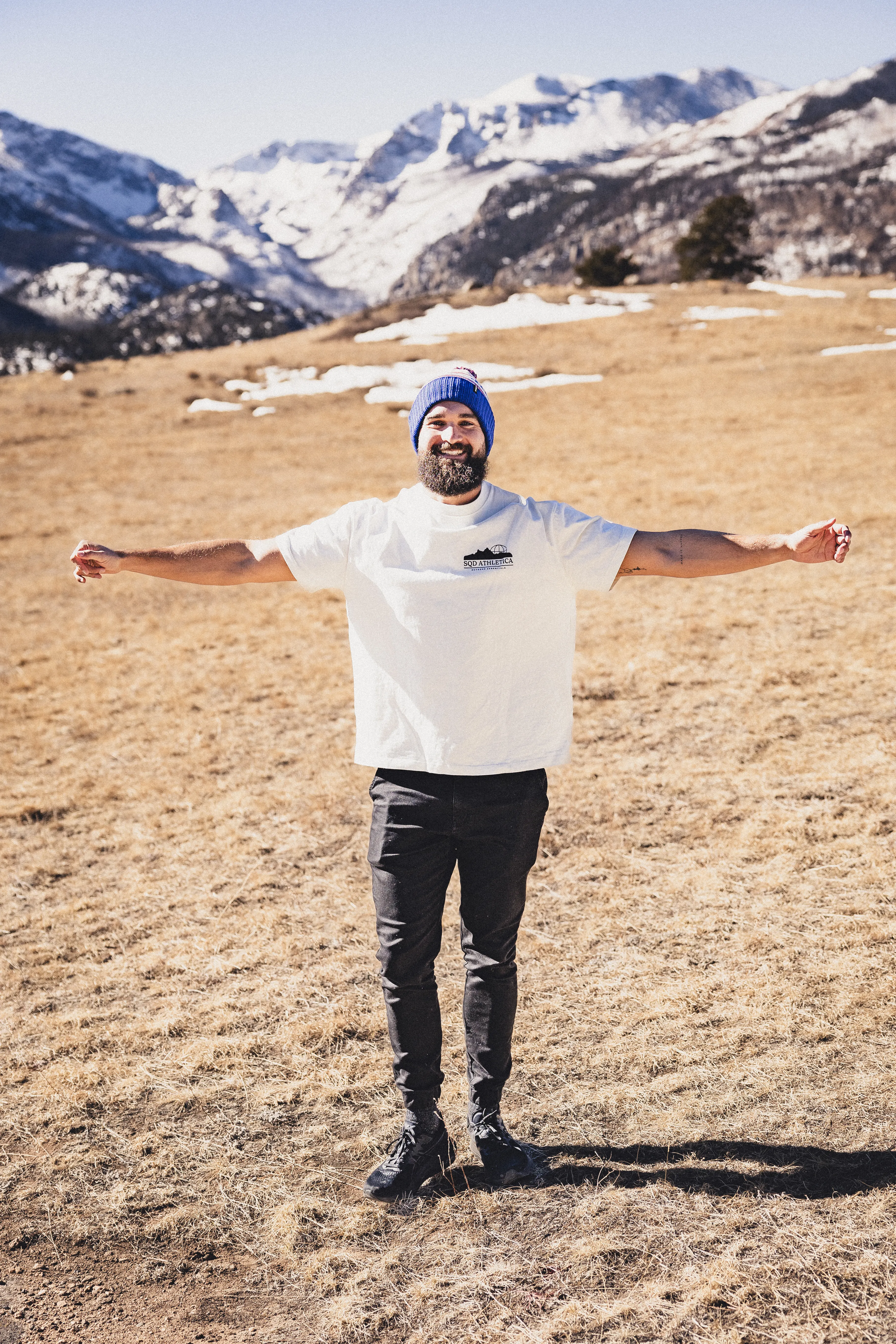 Bearded man wearing a blue beanie and white t-shirt standing with arms outstretched in a dry grassy field with snow-capped mountains in the background.