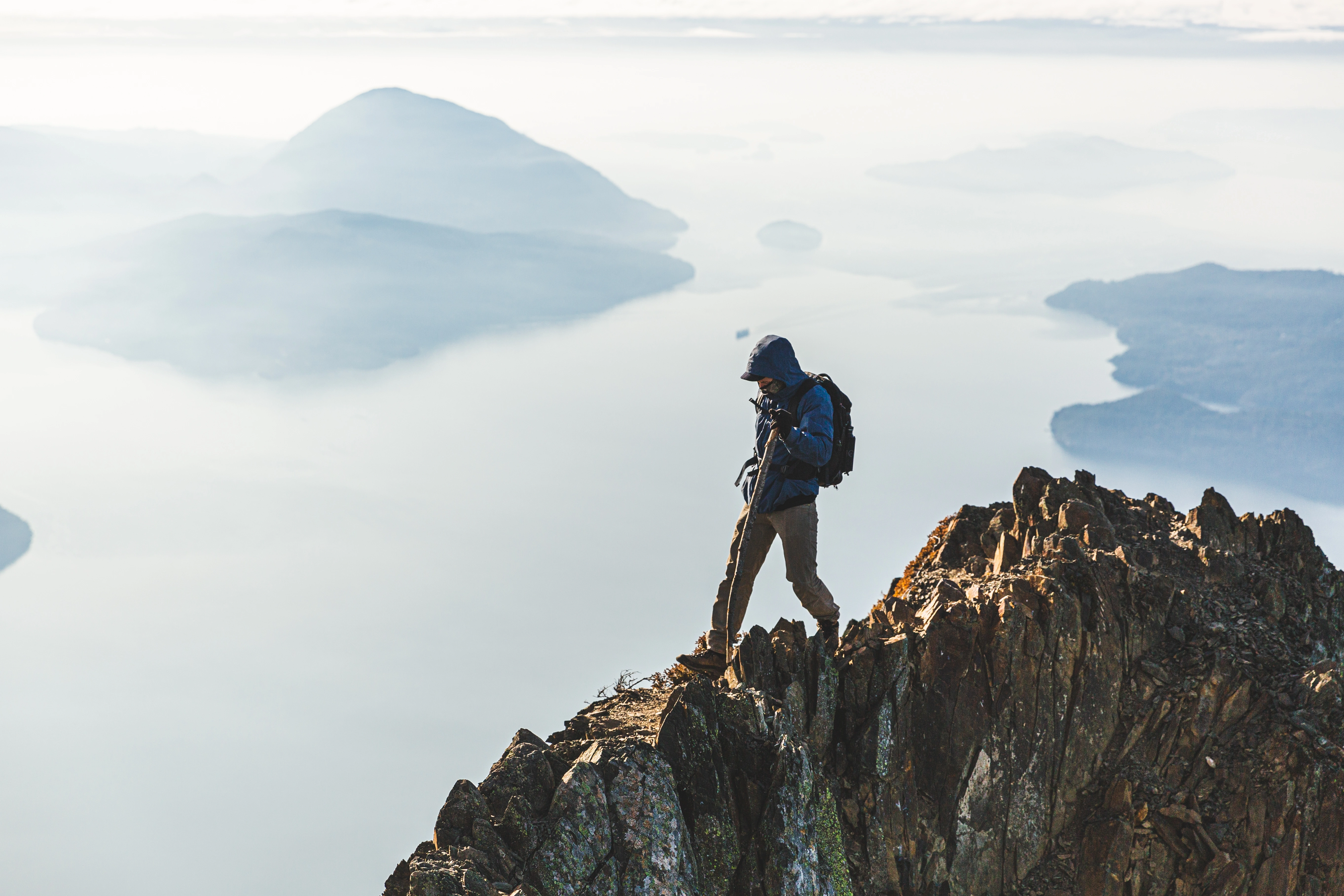 Hiker with a backpack walking on rocky mountain ridge above misty landscape and distant islands.