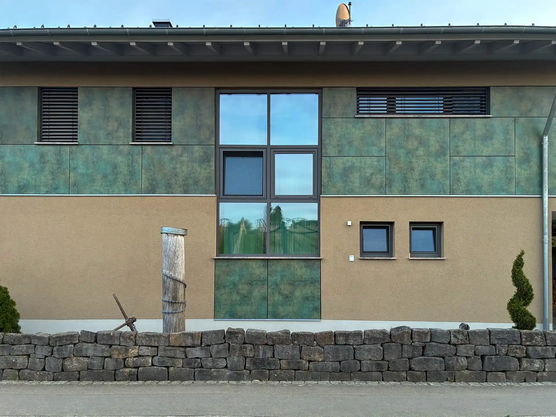 Moderne Hausfassade mit grün-braunen Verkleidungen, großen und kleinen Fenstern sowie einer Steinmauer im Vordergrund.