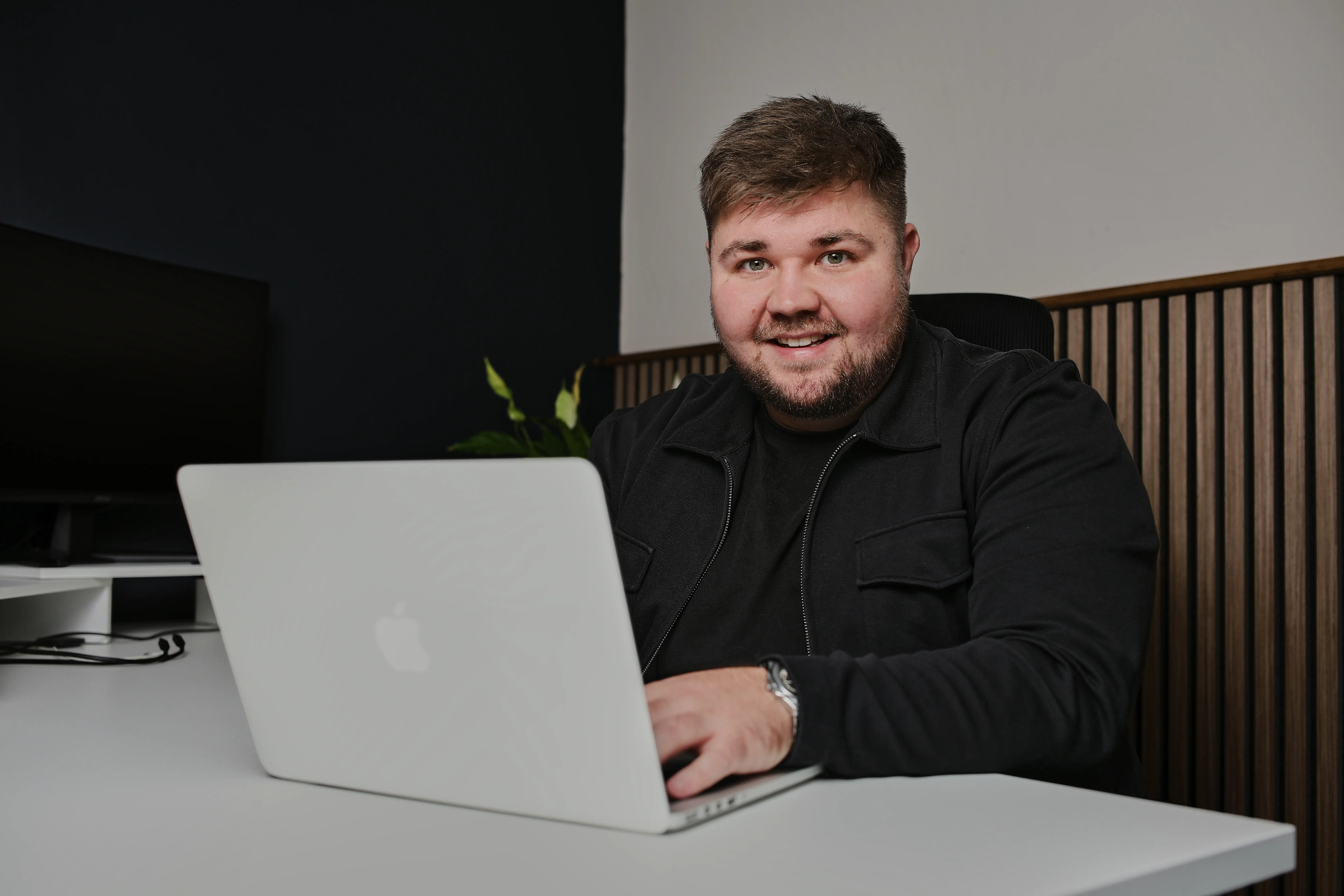 Smiling man with short hair and beard sitting at a white desk, working on a silver Apple laptop in a modern office with dark and light walls and a wooden panel backdrop.