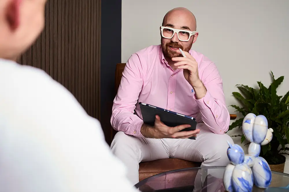 A bald man with a beard and white glasses in a pink shirt and white pants sits on a brown chair, holding a tablet and gesturing with his hand during a conversation with another person whose shoulder is partly visible.