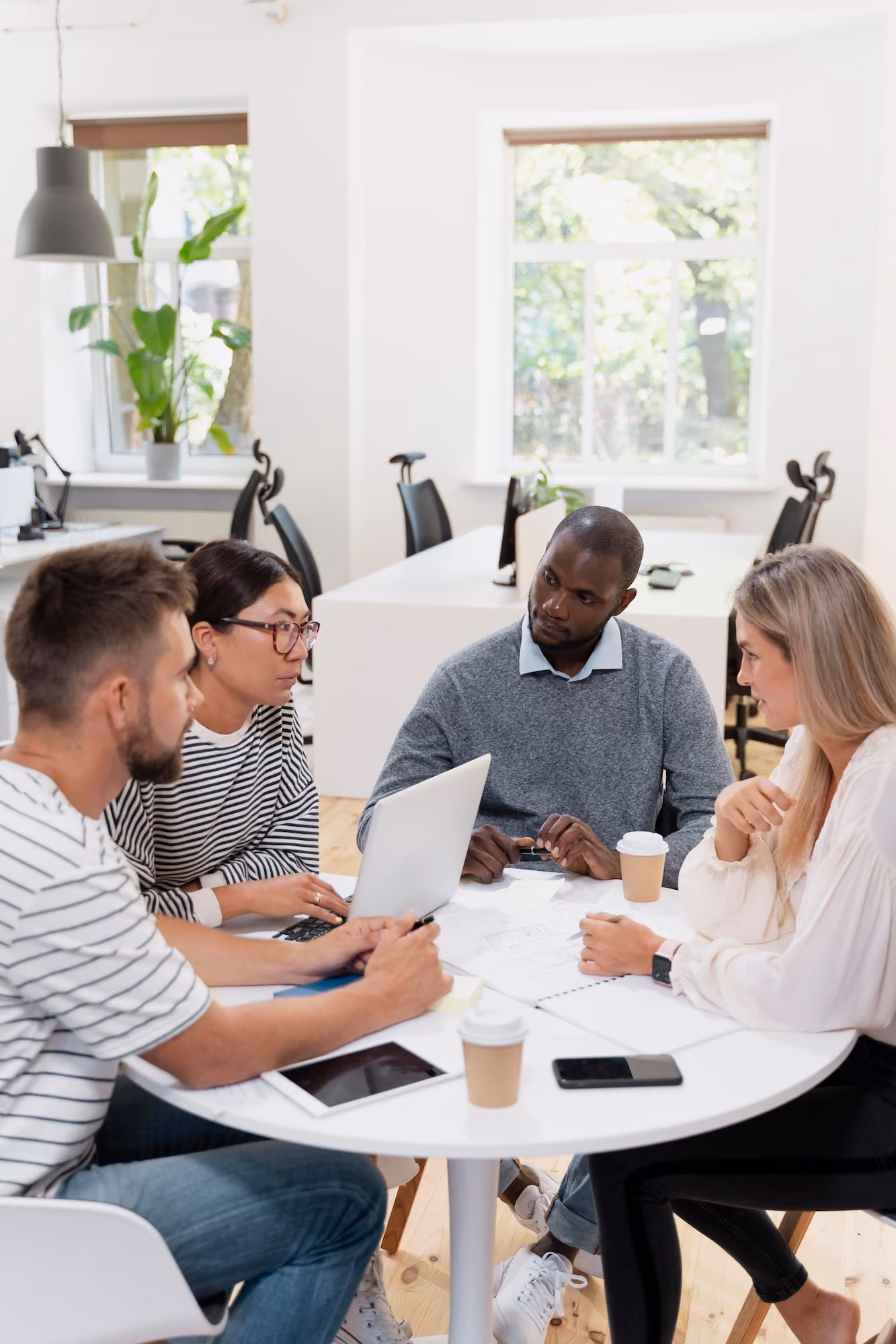 Four young colleagues sitting around a round white table in a bright modern office, engaged in discussion with a laptop, coffee cups, tablet, and smartphone on the table.