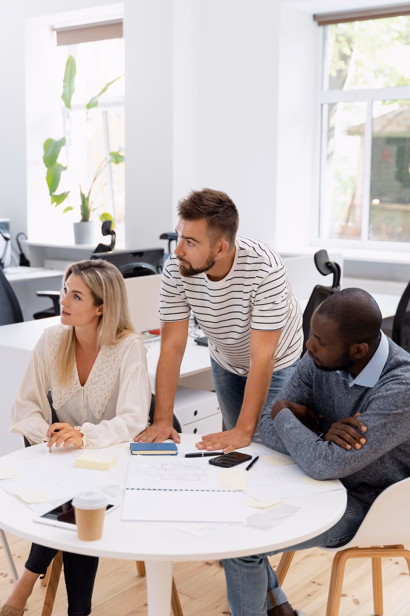 Three colleagues collaborate at a round white table in a bright office; a blonde woman holds a pen, a bearded man in a striped shirt leans on the table, and a man in a grey sweater with crossed arms listens attentively, with papers, notebooks, a smartphone, and a coffee cup on the table.