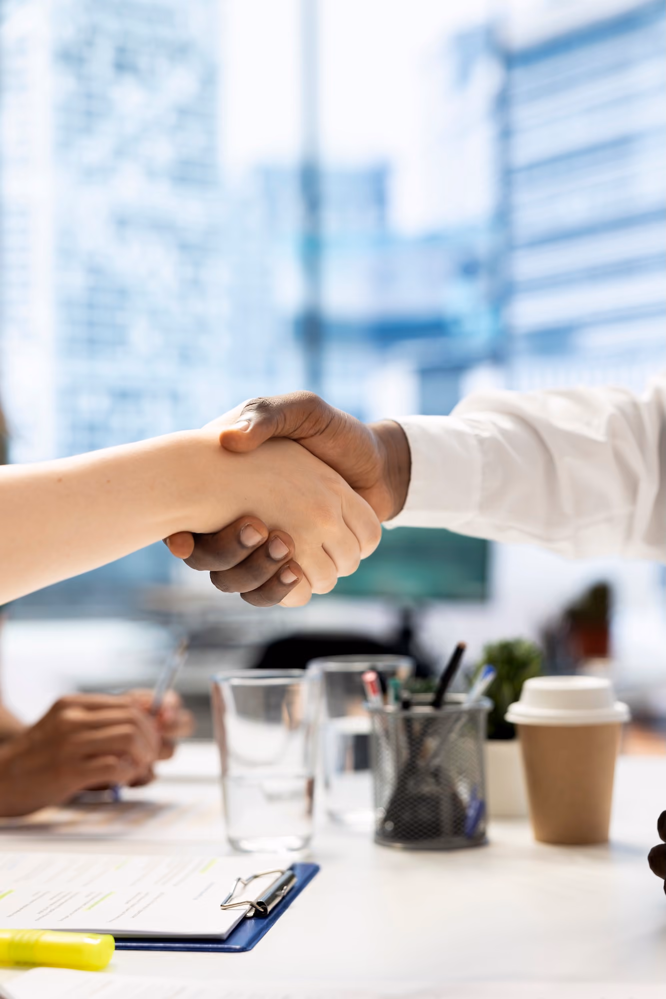 Two businesspeople shaking hands over a desk with office supplies, clipboard, glass of water, and a takeaway coffee cup in a modern office with large windows.