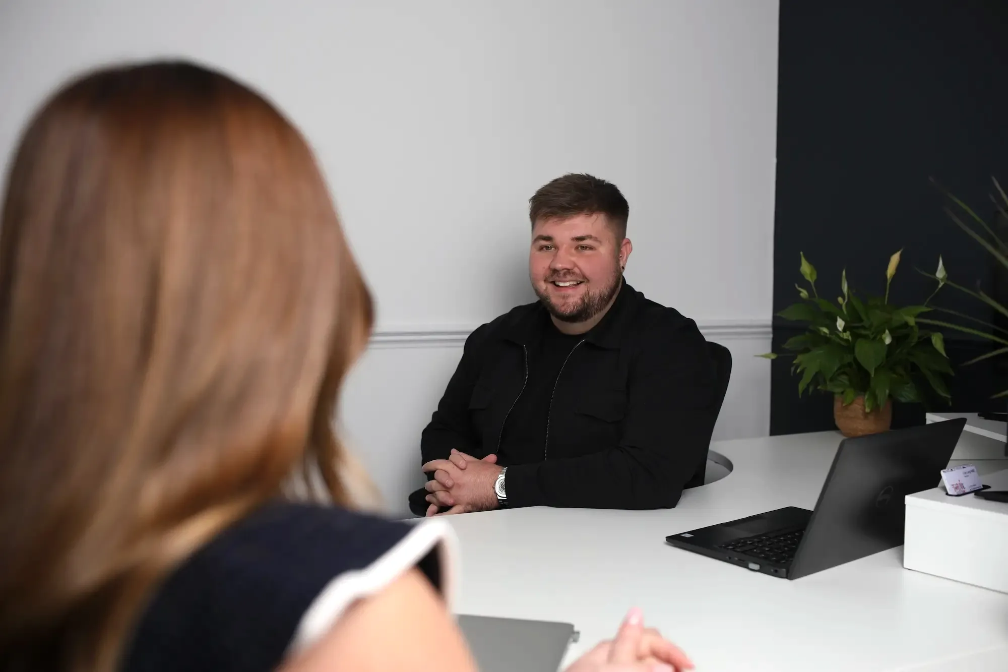 A man smiling and clasping his hands in front of him while sitting at a white desk with a laptop; a woman with long brown hair is seated across from him in an office with light and dark walls and a potted plant in the background.