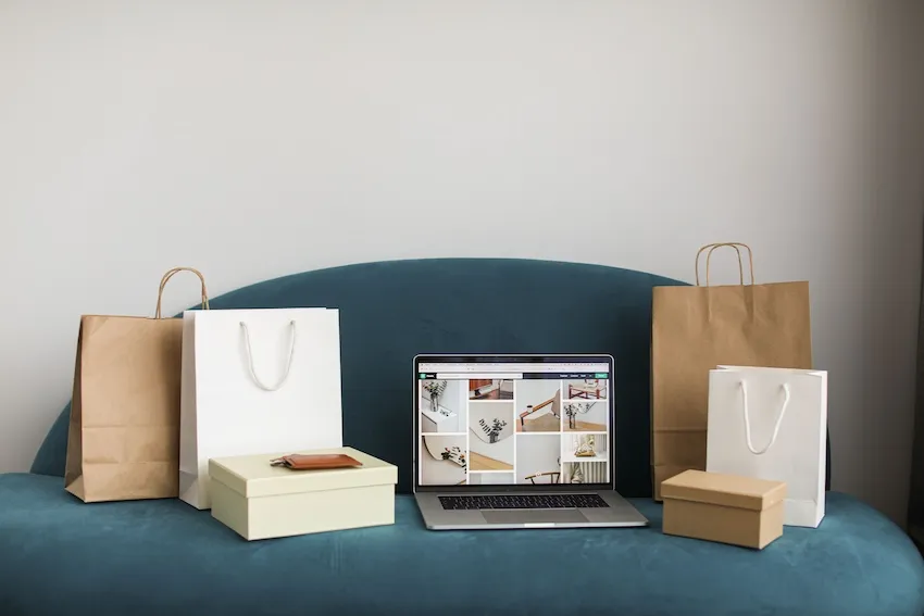 A laptop placed on a blue sofa displaying a collage of interior design photos, flanked by brown and white shopping bags and two closed gift boxes.