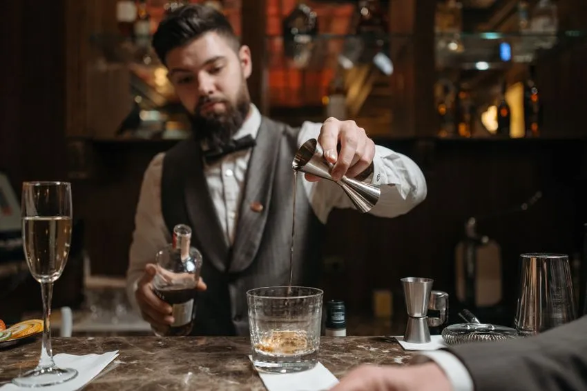 Bearded bartender in a vest and bow tie pouring a dark liquid from a jigger into a glass on a bar counter.