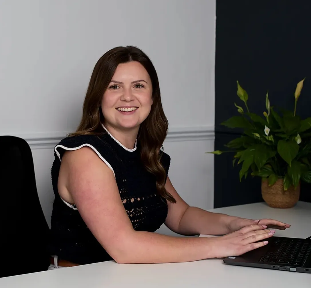 Smiling woman with long brown hair and a black sleeveless crochet top sitting at a desk working on a laptop, with a plant and wood paneling in the background.