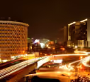 Night cityscape with blurred light trails from vehicles on a busy curved road and illuminated buildings.