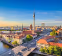 Cityscape with TV tower, several buildings, and streets under a blue sky at sunset.