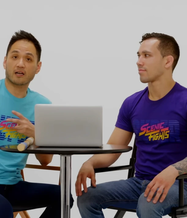 Two men sitting at a round table with a laptop; both wear Scenic Fights t-shirts in blue and purple colors.