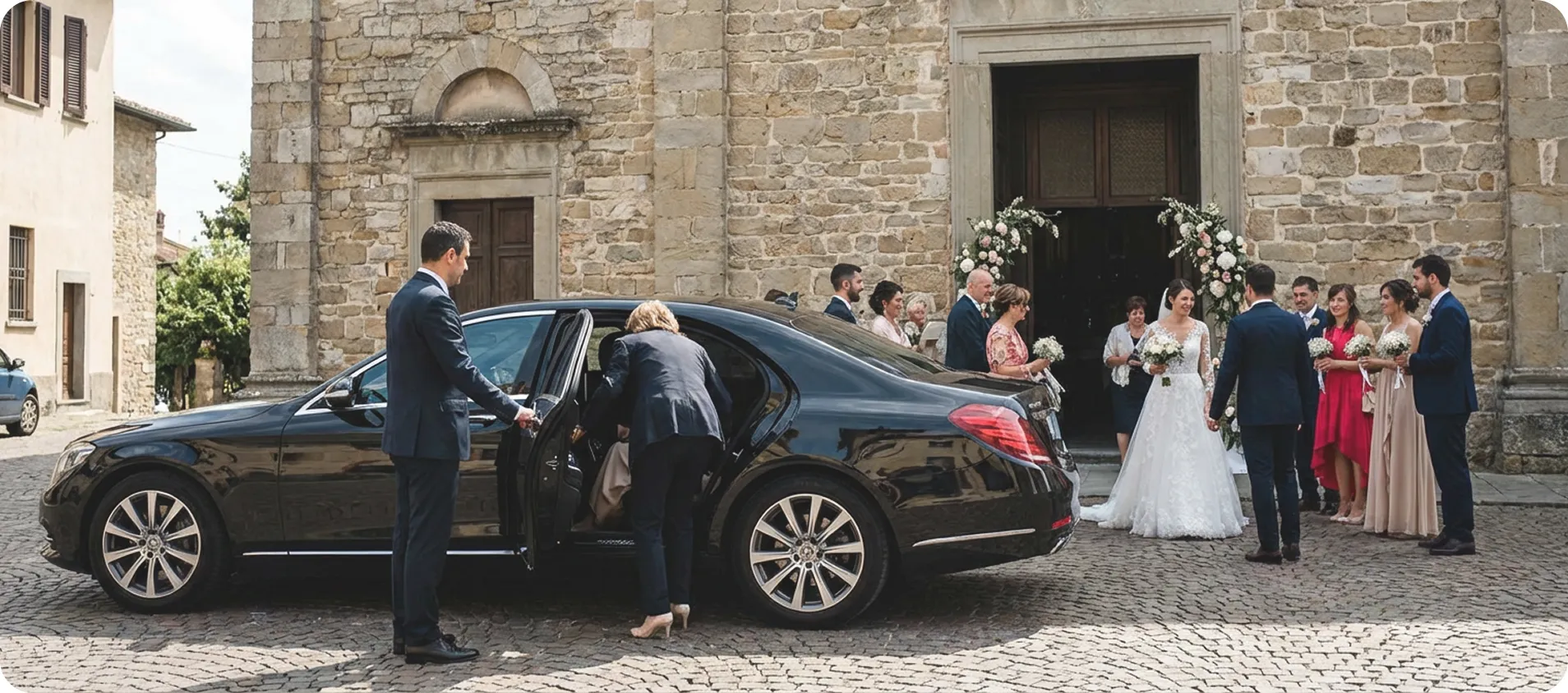 A bride and groom with their wedding party stand outside a stone building decorated with flower arches, while a black luxury car is parked nearby with a woman getting in and a man holding the door.