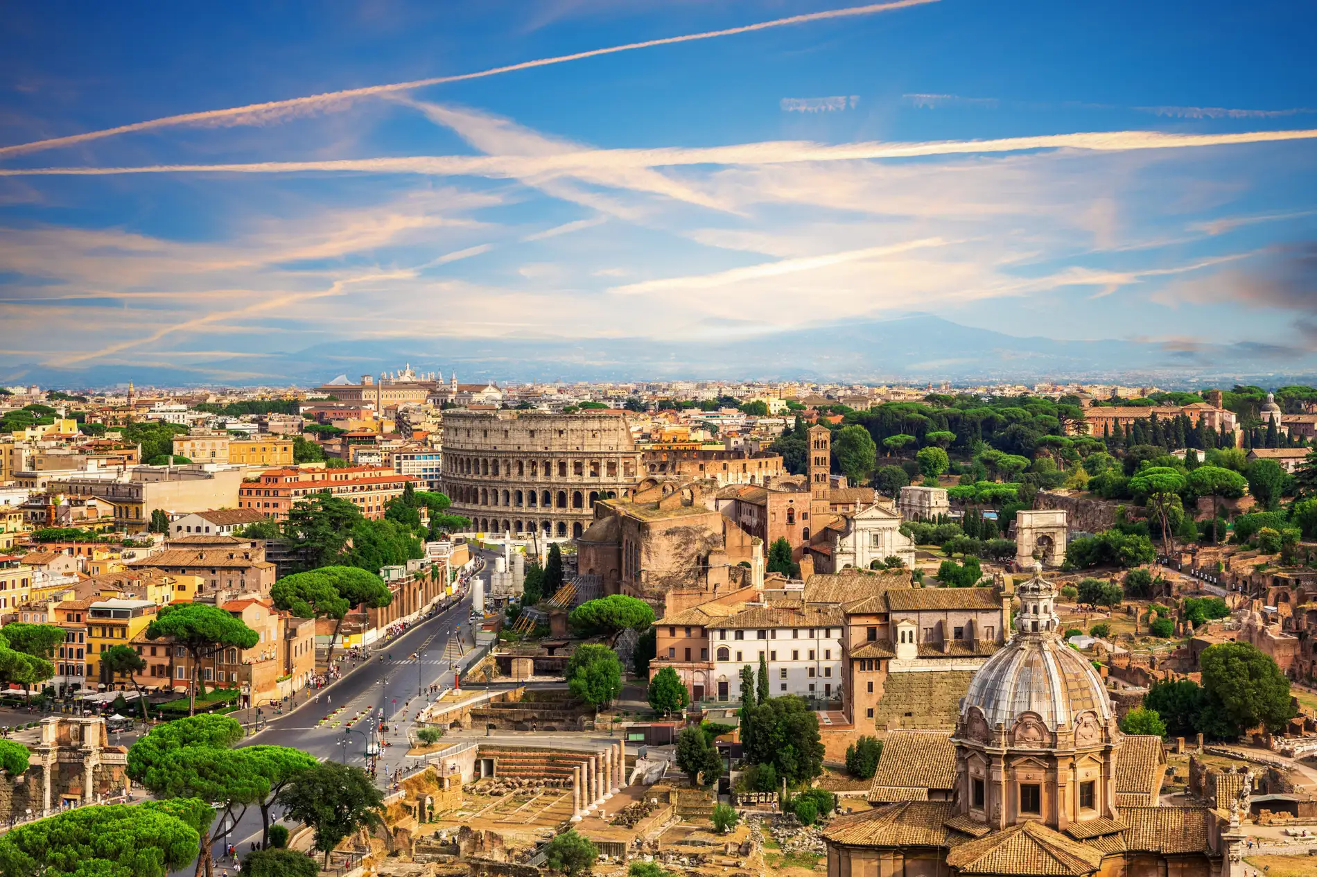 Panoramic view of Rome featuring the Colosseum, ancient ruins, historic buildings, and green trees under a blue sky with contrails.