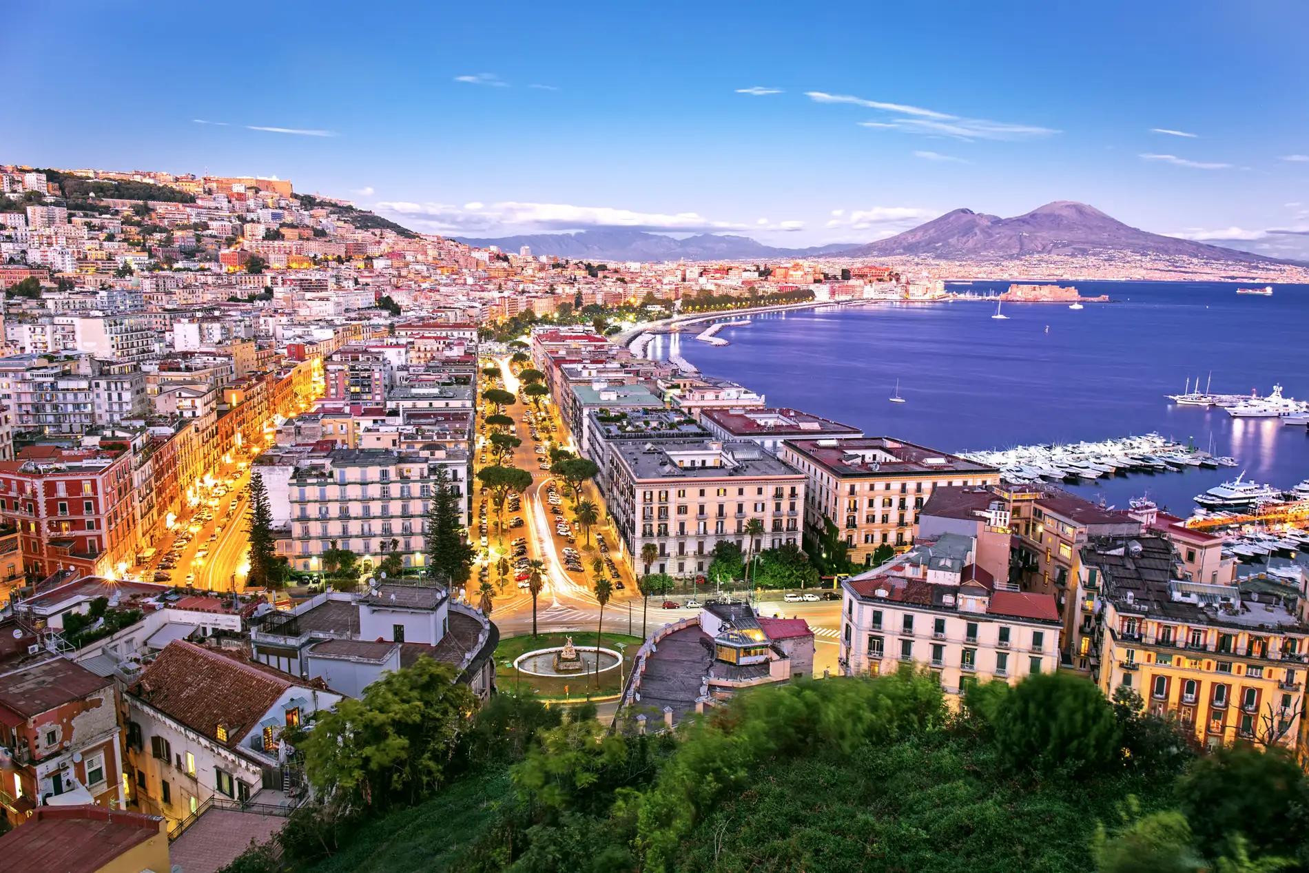 Panoramic view of Naples, Italy at dusk with illuminated streets, coastal buildings, marina, and Mount Vesuvius in the background.