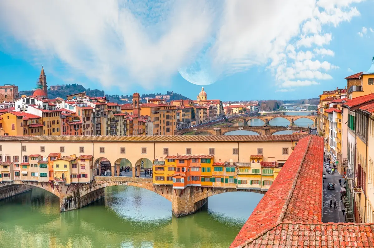 Panoramic view of the Ponte Vecchio bridge spanning the Arno River in Florence, Italy, under a partly cloudy sky.