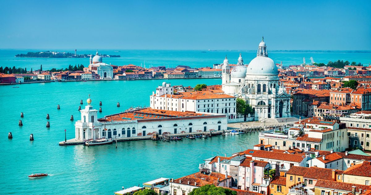 Aerial view of Venice, Italy, showing the turquoise waters of the lagoon, historic buildings with red roofs, and the prominent white domes of Basilica di Santa Maria della Salute.
