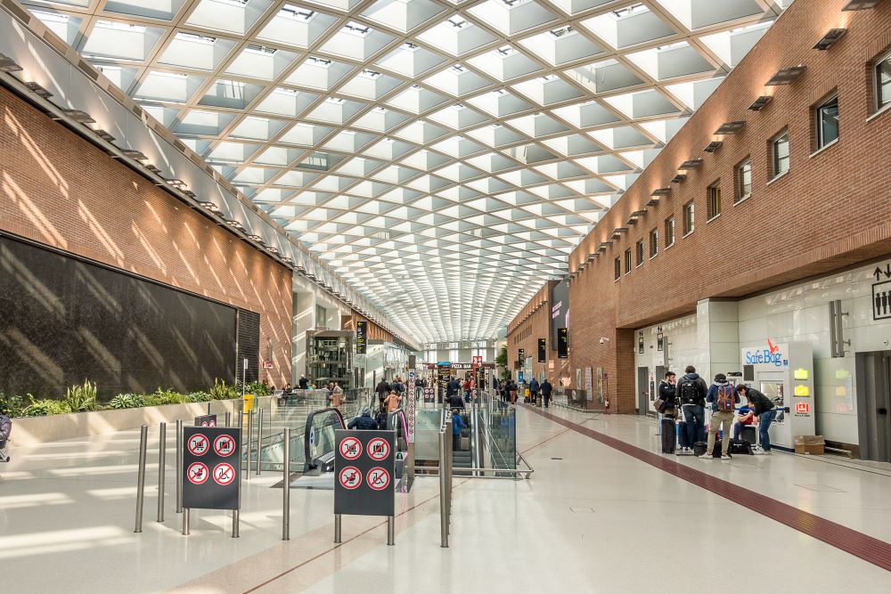 Spacious, modern airport terminal with a glass ceiling, brick walls, escalators, and travelers with luggage.