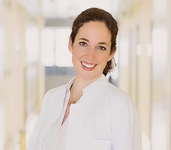 Smiling woman with dark hair wearing a white lab coat in a bright hallway.