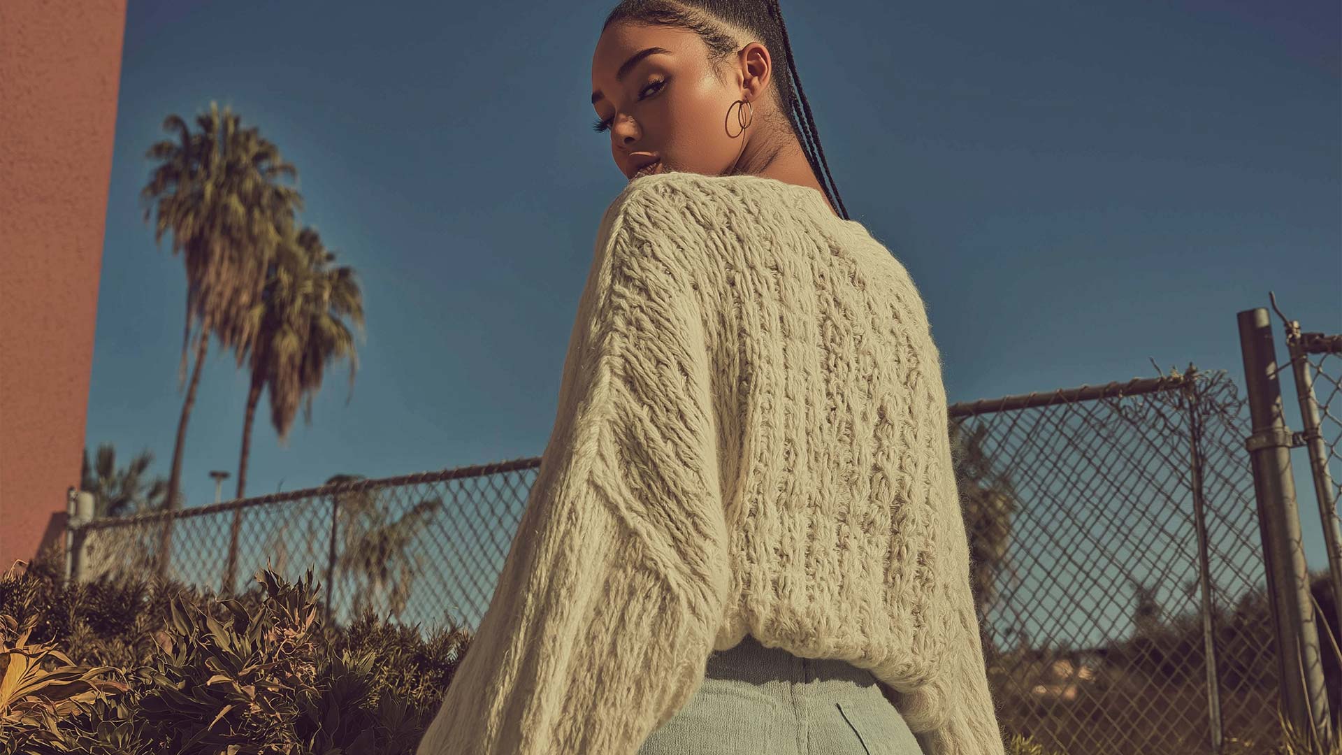 Woman seen from behind wearing a loose knit sweater and fitted pants, looking over her shoulder outdoors near a chain-link fence and palm trees under a clear blue sky.