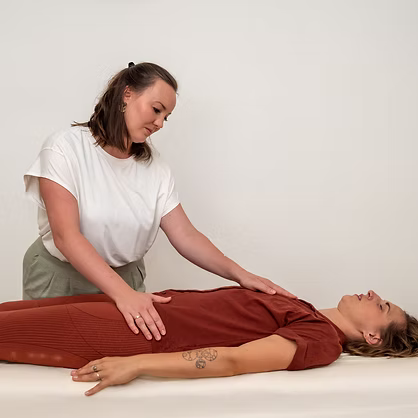 Sarah Lux bei einer Einheit integrative Körperarbeit auf der Liege. Sie berührt sanft den Bauch einer liegenden Frau in braunem Kleid.