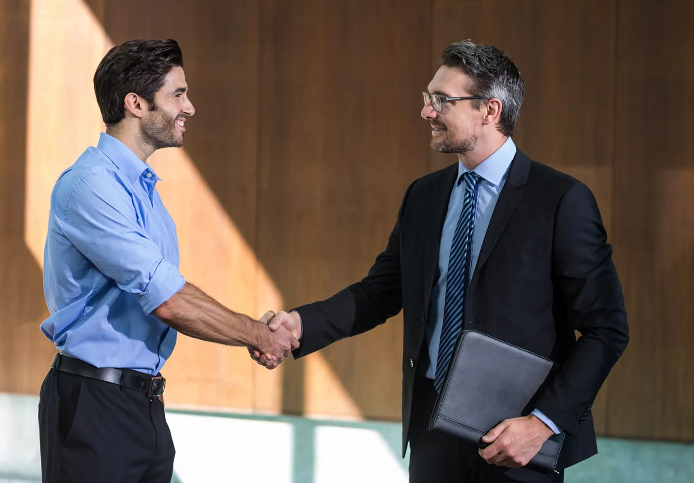 Two men smiling and shaking hands indoors, one in a blue shirt and the other in a black suit holding a black portfolio.