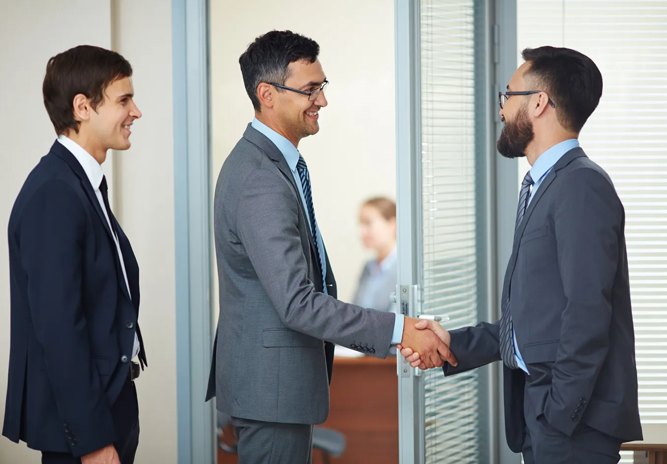Two men in suits and glasses shaking hands in an office doorway while a third man smiles nearby.