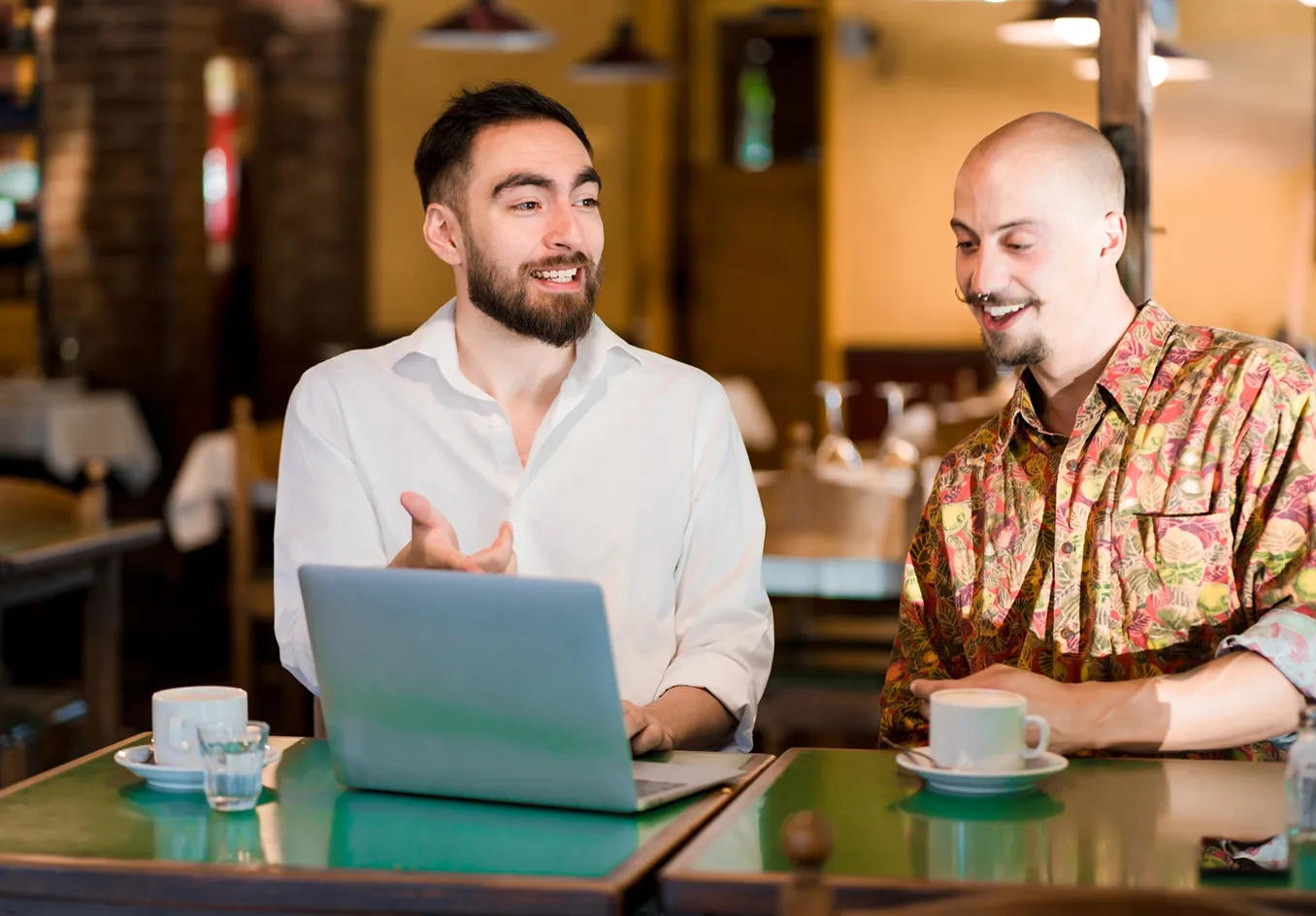 Two men sitting at a table in a cafe, one with a laptop gesturing while the other listens with a cup of coffee.