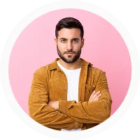 Man with dark hair and beard, wearing a brown jacket and white t-shirt, standing with arms crossed against a pink background.