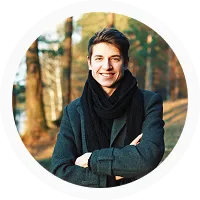 Young man with dark hair wearing a black scarf and coat, smiling with arms crossed outdoors in a wooded area.