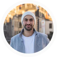 Smiling man with beard wearing a white knit hat and light gray jacket standing outdoors with a blurred cityscape background.