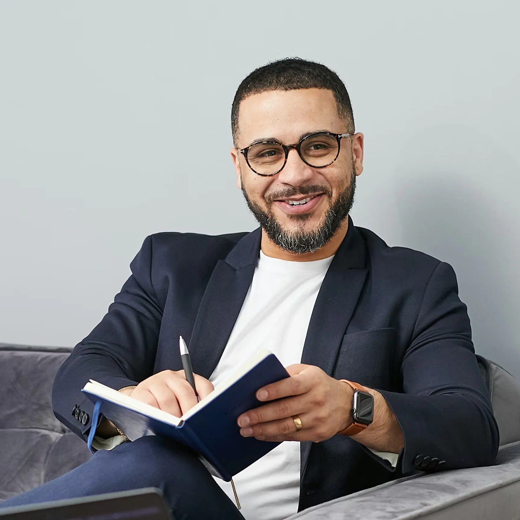 Smiling man with glasses and beard wearing a dark blazer writing in a blue notebook while sitting on a gray couch.