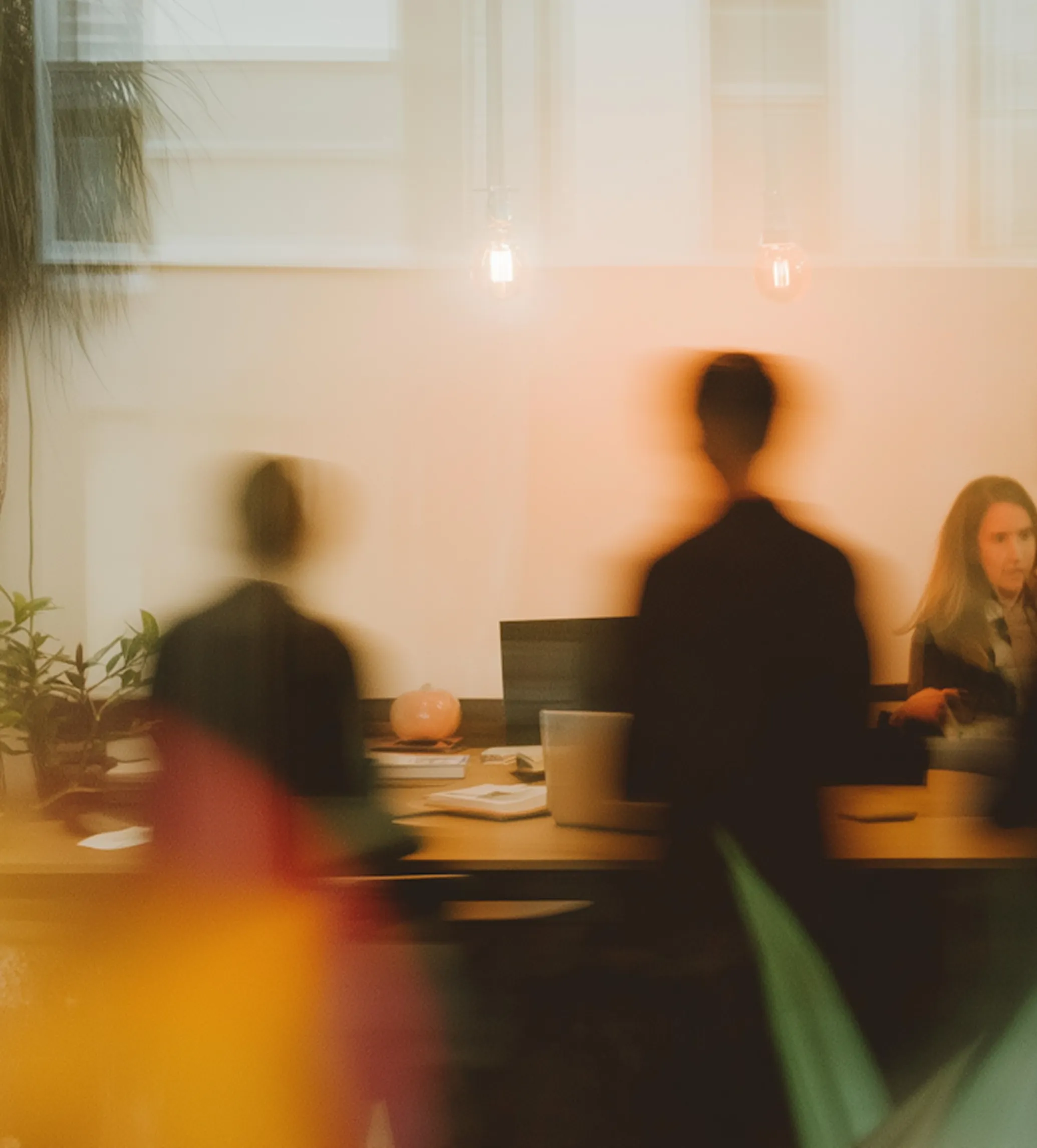 Blurred silhouettes of people sitting at a wooden table with laptops and books in a softly lit room.