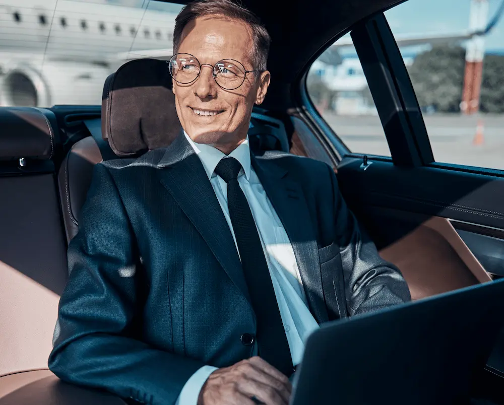Homme d'affaires souriant portant un costume et des lunettes, assis à l'arrière d'une voiture de luxe avec un ordinateur portable.
